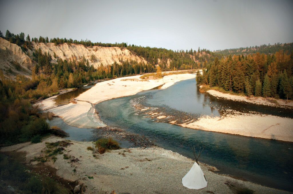 A white teepee next to the riverbank of the St. Mary River